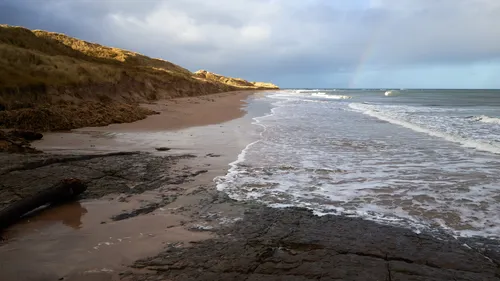 This landscape photograph features a Northumbrian winter beach during the early afternoon, with soft light illuminating the dunes and coastline. The image depicts the sandy beach and tidal shoreline along the Northumberland coast near Seahouses and Bamburgh in England, United Kingdom. Waves from the sea gently lap the shore, while a faint rainbow can be seen in the sky above the coastal landscape. The photograph captures the natural beauty of the Northumberland coast, emphasising the wintry atmosphere and the expanse of beach and sea typical of this region.